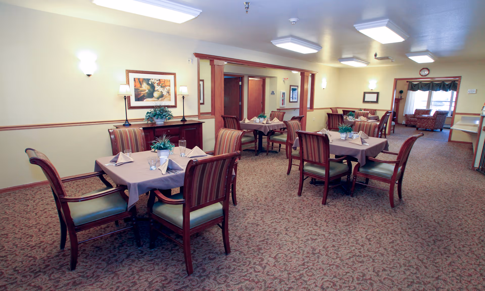 Dining room with multiple tables set for meals and upholstered chairs in a senior living facility.