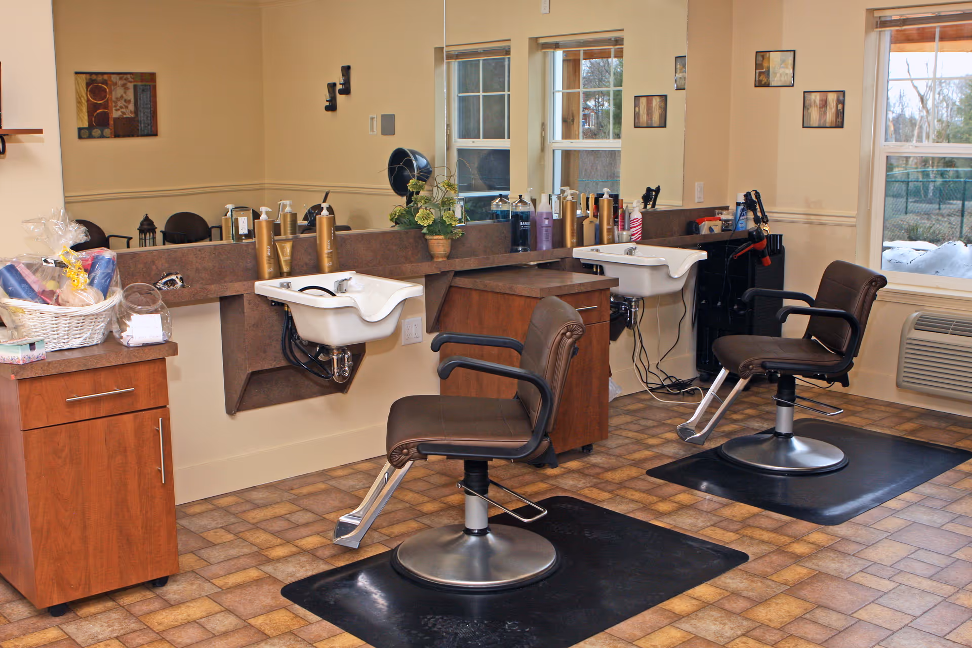 Interior view of a hair salon area with two brown salon chairs in front of two white wash basins. The room has a large mirror, various hair care products on the counter, a wooden cabinet with a basket of towels and supplies, and windows showing an outdoor view.