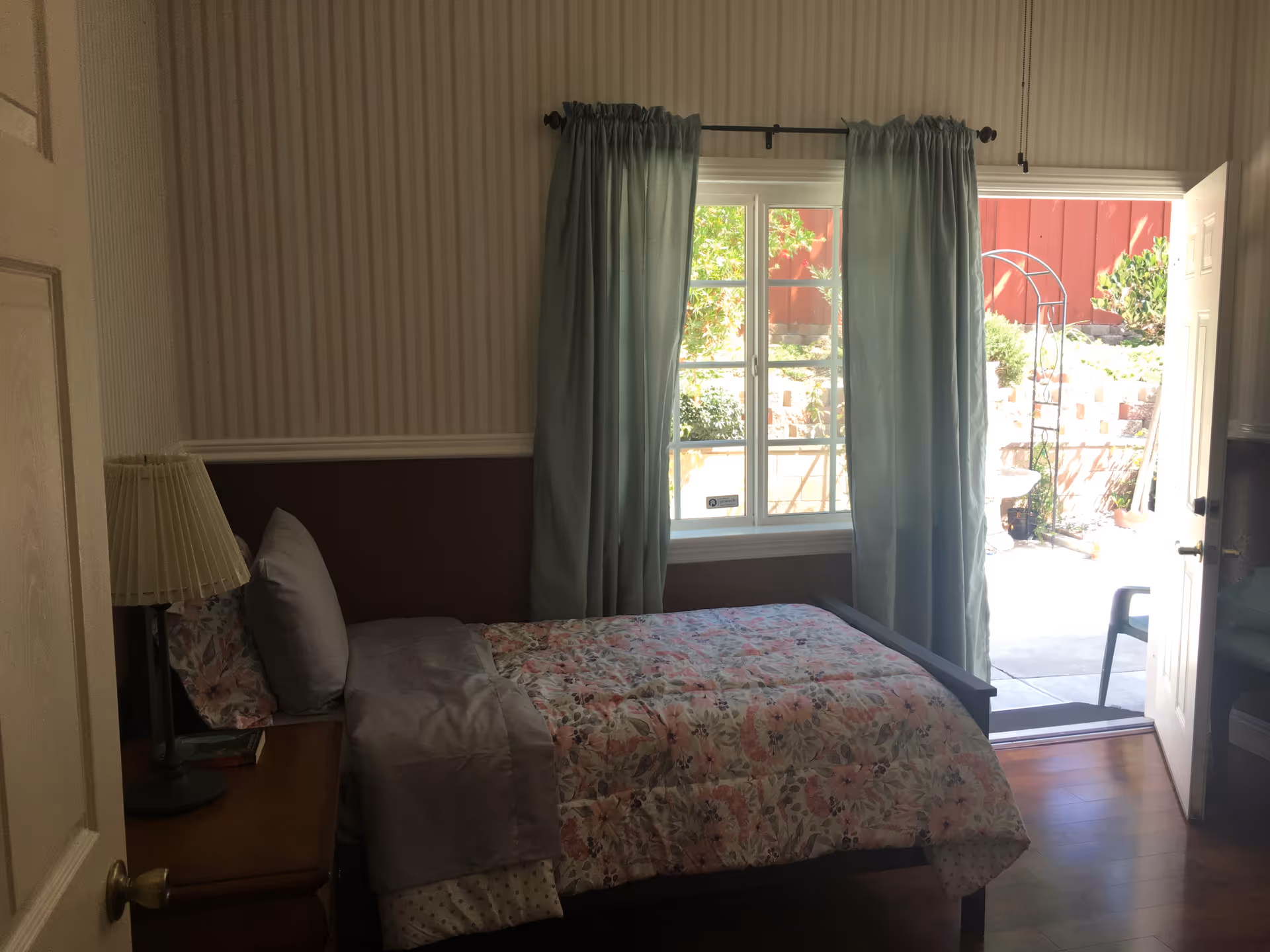 Cozy bedroom with floral bedspread, bedside lamp, window, and open door to patio.
