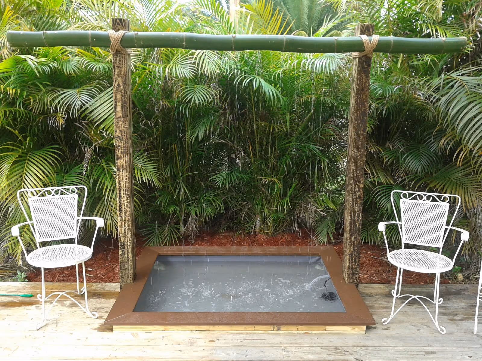 An outdoor water feature with a rectangular basin filled with water, surrounded by a wooden frame. Water is falling from a horizontal bamboo pipe supported by two vertical wooden posts. Two white metal chairs with armrests are positioned on either side of the water feature. Lush green tropical plants form the background.