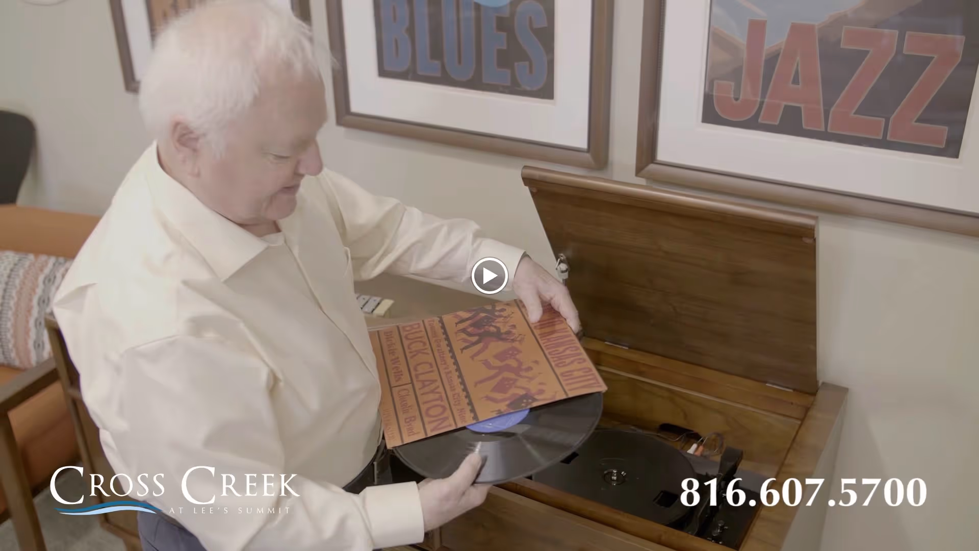 An elderly man with white hair is placing a vinyl record on a wooden record player in a cozy room. Behind him, there are framed posters on the wall with the words 'BLUES' and 'JAZZ'. A patterned cushion is visible on a chair in the background.