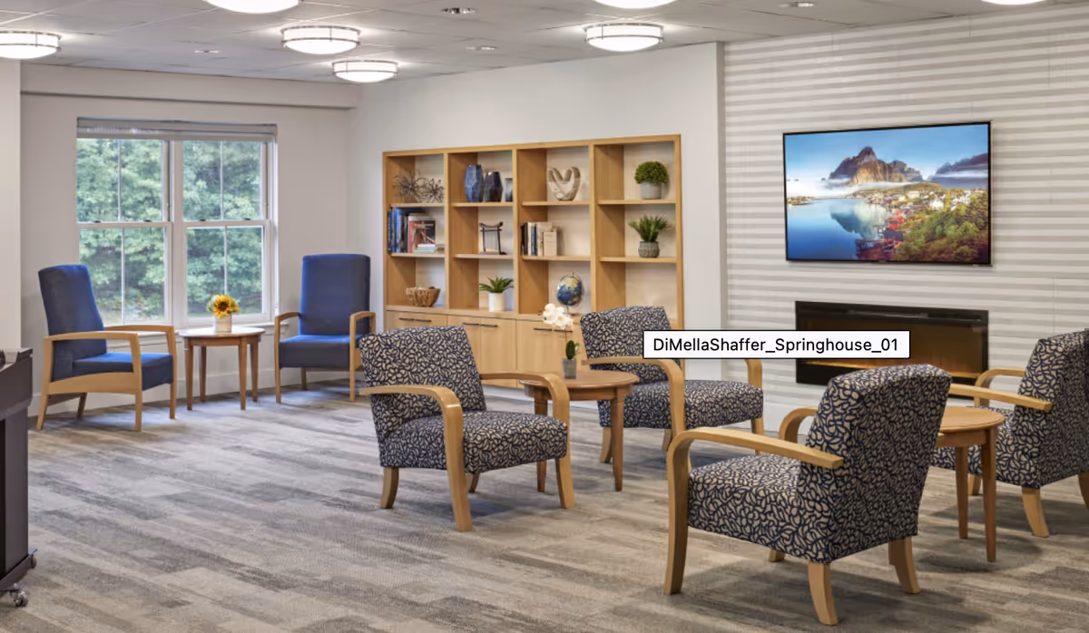 A senior living facility common area with several patterned armchairs and wooden side tables arranged on a carpeted floor. There is a large window with a view of greenery outside, a wooden shelving unit with decorative items and books, and a wall-mounted flat-screen TV above a modern fireplace.