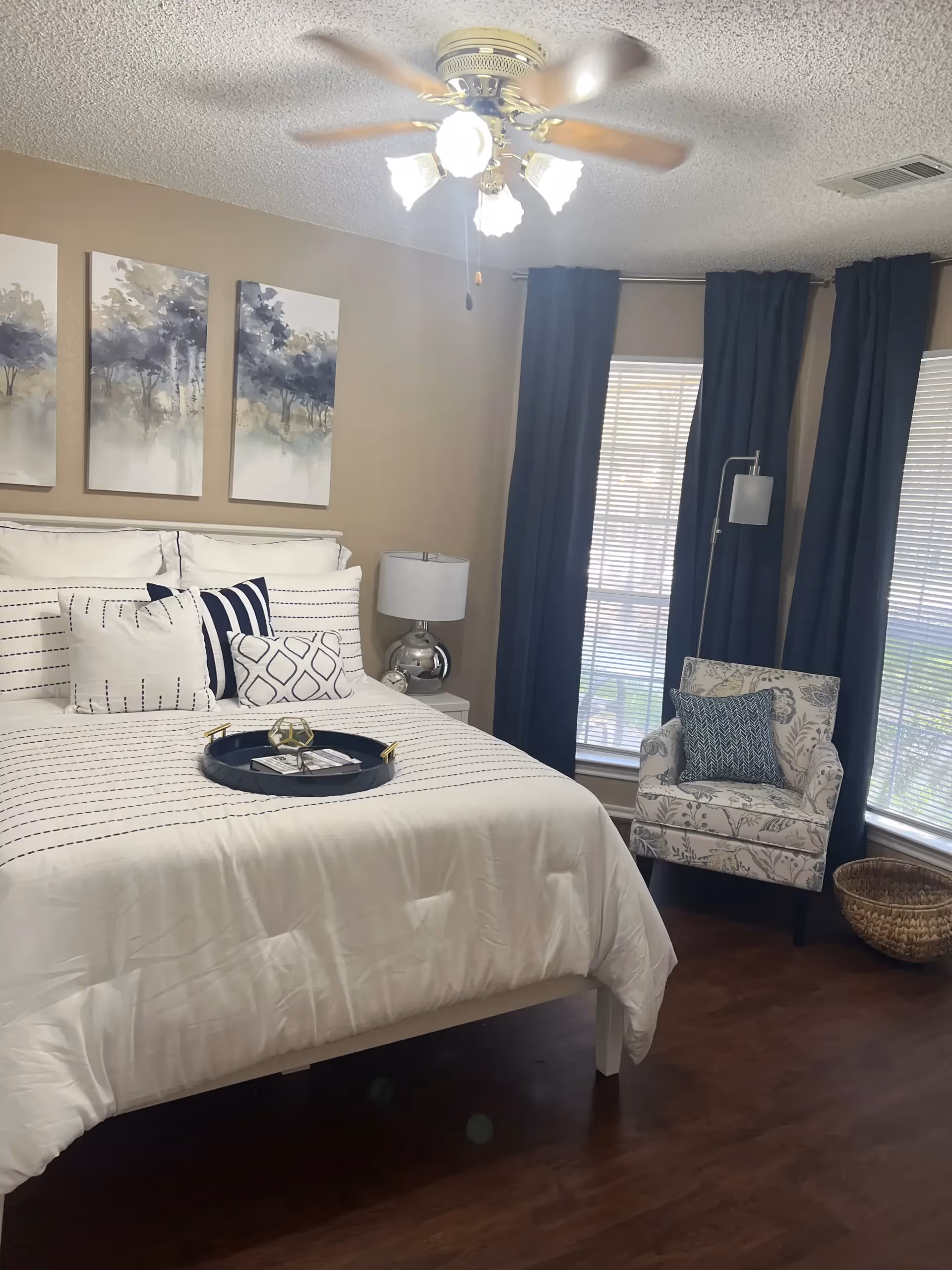 Neatly made bedroom with a white bed and decorative pillows, a nightstand with a lamp, an upholstered armchair by tall windows with dark curtains, and a ceiling fan.
