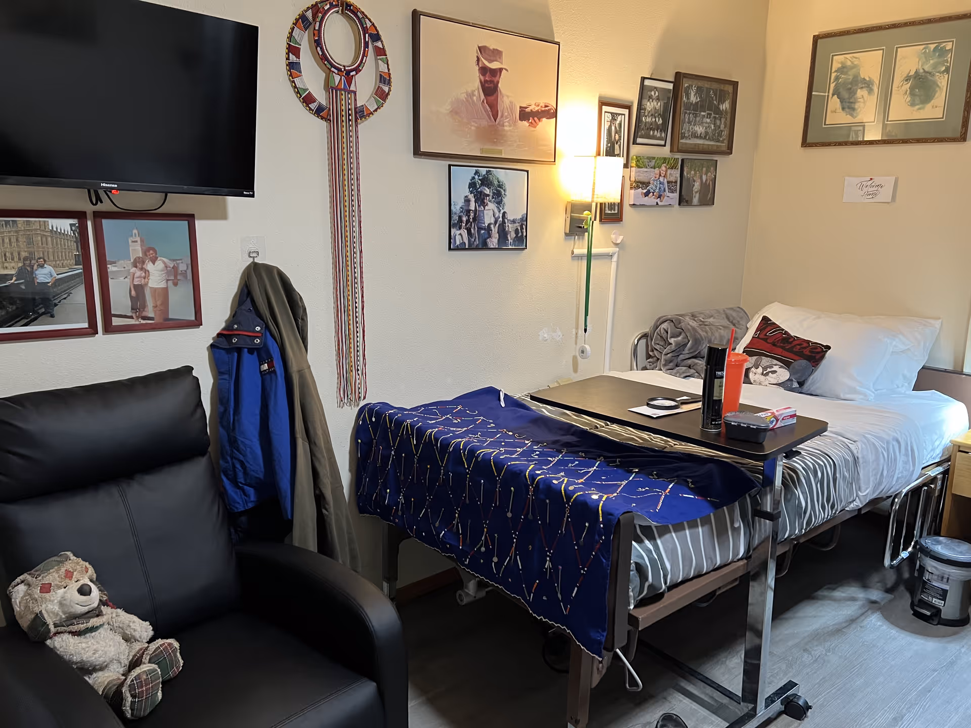Small furnished bedroom in an adult family home with a hospital-style bed, overbed table, recliner and framed photos on the walls.