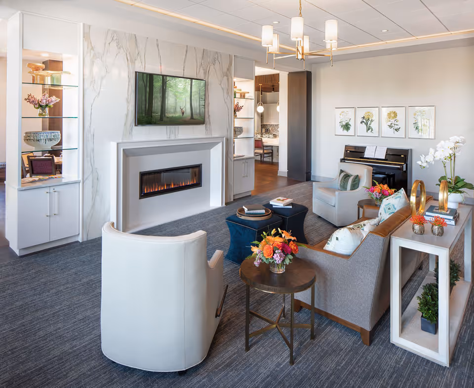 A modern and cozy living room in a senior living facility featuring a white armchair, a gray sofa with decorative pillows, a small round side table with a flower arrangement, and a fireplace with a mounted TV above it displaying a forest scene. The room also includes built-in shelves with decorative items, a piano against the wall with sheet music, botanical artwork, and a chandelier hanging from the ceiling.