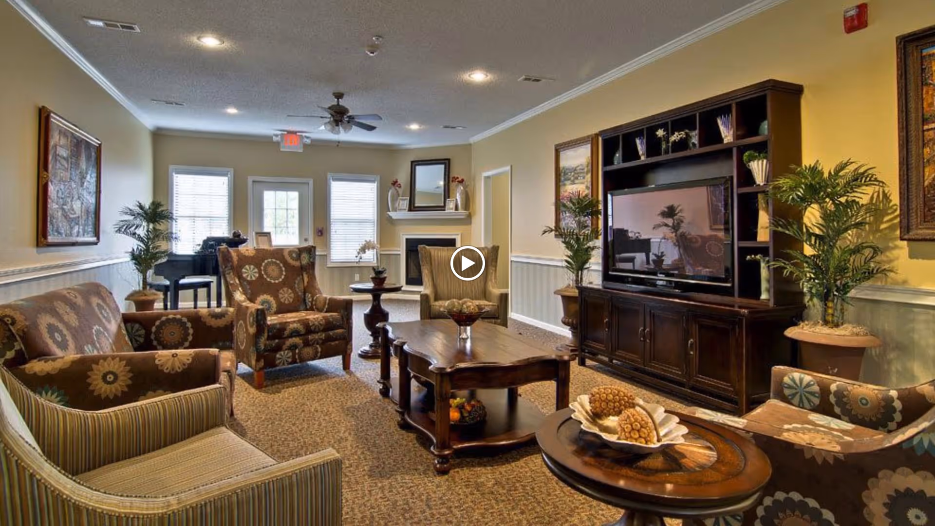 Comfortable common living room with patterned armchairs, coffee tables, a large TV cabinet, potted plants and a piano in the background.