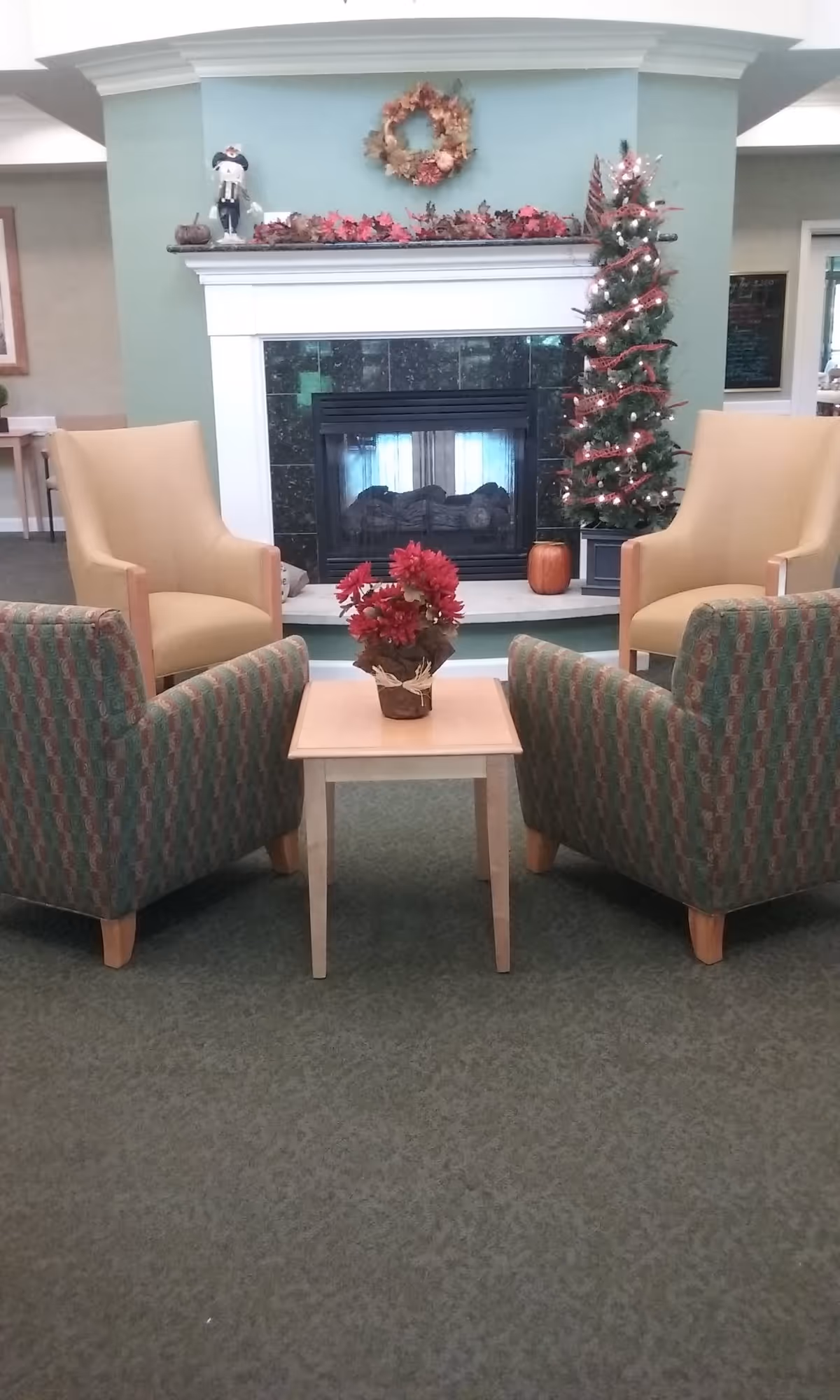 A cozy seating area in a senior living community featuring two beige armchairs and two patterned armchairs arranged around a small wooden table with a red flower centerpiece. Behind the seating area is a fireplace decorated with autumn leaves, a wreath, a small scarecrow figure, and a small decorated Christmas tree with lights and red ribbon.