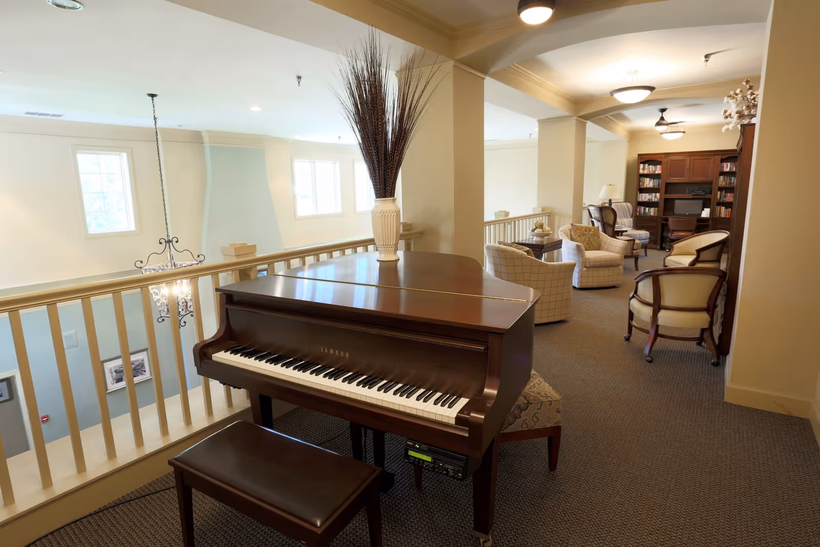 A cozy interior common area featuring a Yamaha grand piano with a vase of tall decorative branches on top. The space includes several upholstered chairs arranged near a wooden bookshelf filled with books and a computer desk. The area is well-lit with ceiling lights and natural light from windows.