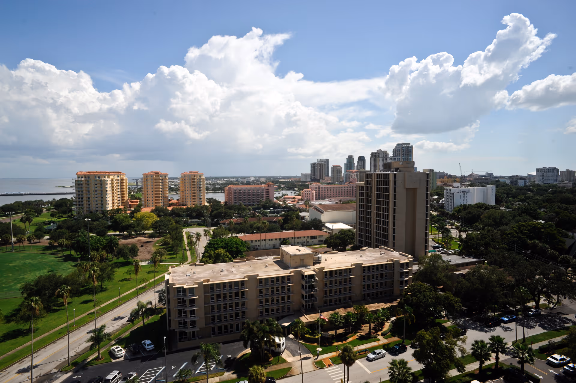 Aerial view of a cityscape featuring multiple mid-rise and high-rise buildings, green spaces, and a body of water in the background under a partly cloudy sky.