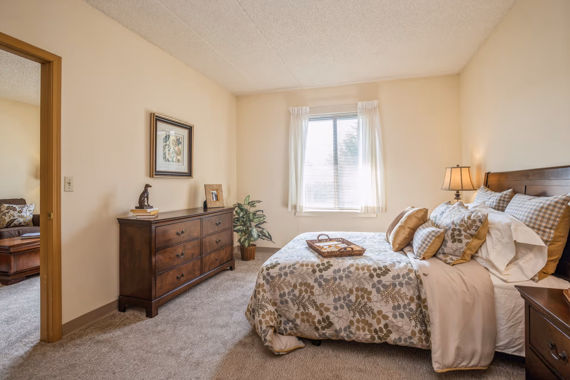 A cozy bedroom with a large bed covered in floral patterned bedding and multiple pillows. A wooden dresser with decorative items and a framed picture hangs above it. A window with white curtains lets in natural light. The room has beige walls and carpeted floor. An open doorway leads to another room with a sofa and coffee table.