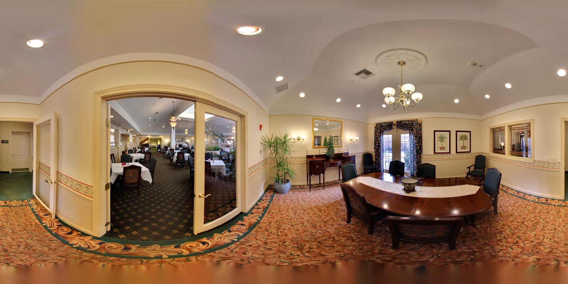 A formal dining room with a large wooden oval table surrounded by six chairs. The room features a chandelier hanging from a decorative ceiling medallion, a sideboard with a mirror above it, framed botanical artwork on the walls, and a potted plant in the corner. Double glass doors lead to another dining area with multiple tables and chairs covered with white tablecloths. The carpet has an ornate floral pattern, and the walls are painted a soft yellow with a decorative border.