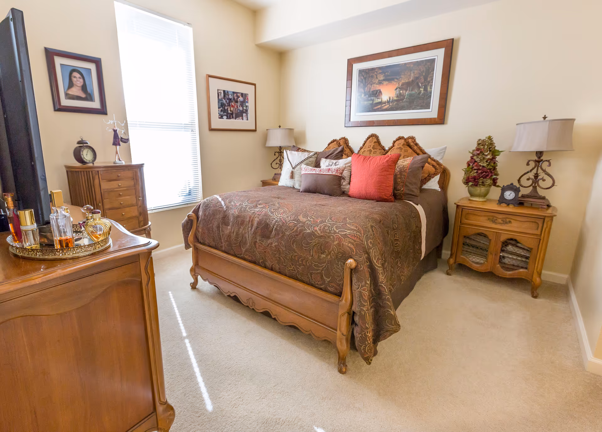 A cozy bedroom with a wooden bed frame and a brown patterned bedspread adorned with multiple pillows. The room has beige walls and carpeted floor. There is a wooden dresser with perfume bottles on a tray, a wooden chest of drawers near a window with blinds, and a wooden nightstand with a lamp, clock, and decorative plant. Two framed pictures hang on the walls.