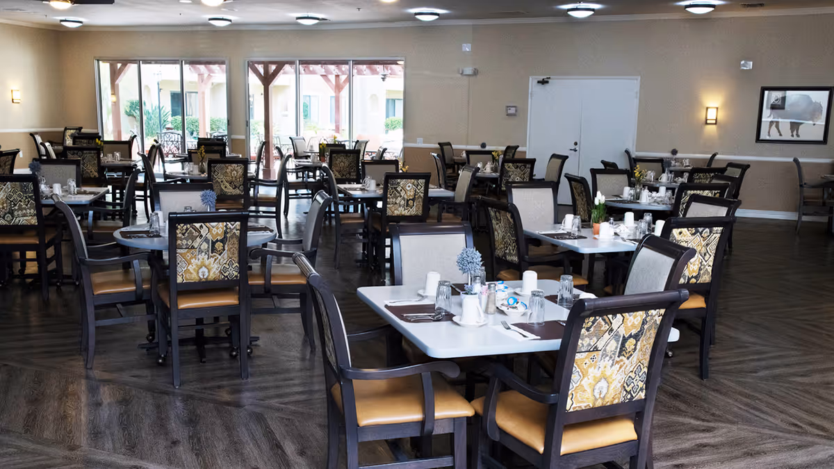 A spacious dining room with multiple tables and chairs arranged neatly. Each table is set with placemats, cups, glasses, and small flower vases. Large windows in the background let in natural light and provide a view of an outdoor patio area.