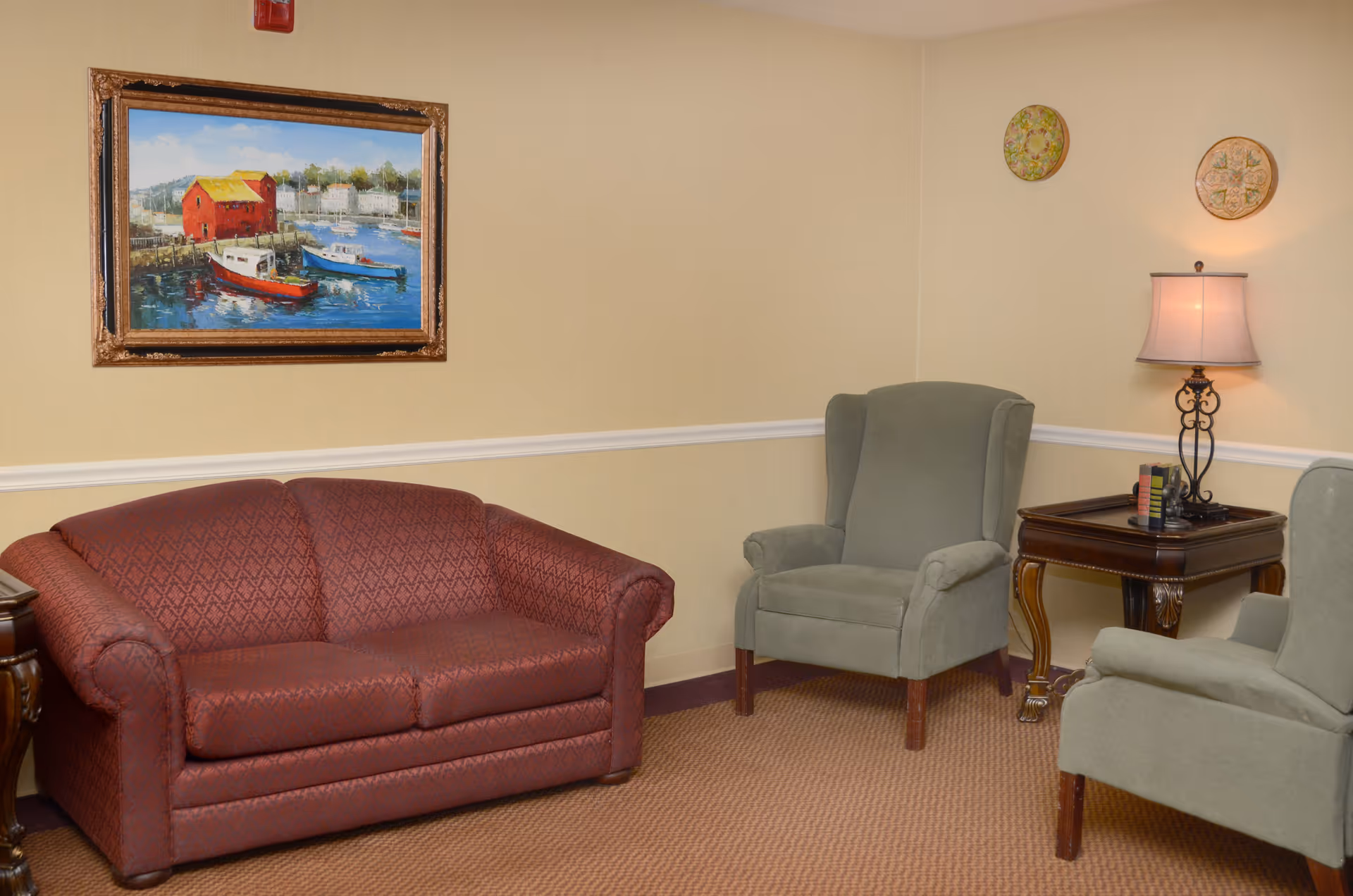 A cozy sitting area with a maroon patterned loveseat, two green upholstered armchairs, a wooden side table with a decorative lamp, and a framed painting of boats on the water hanging on a beige wall.