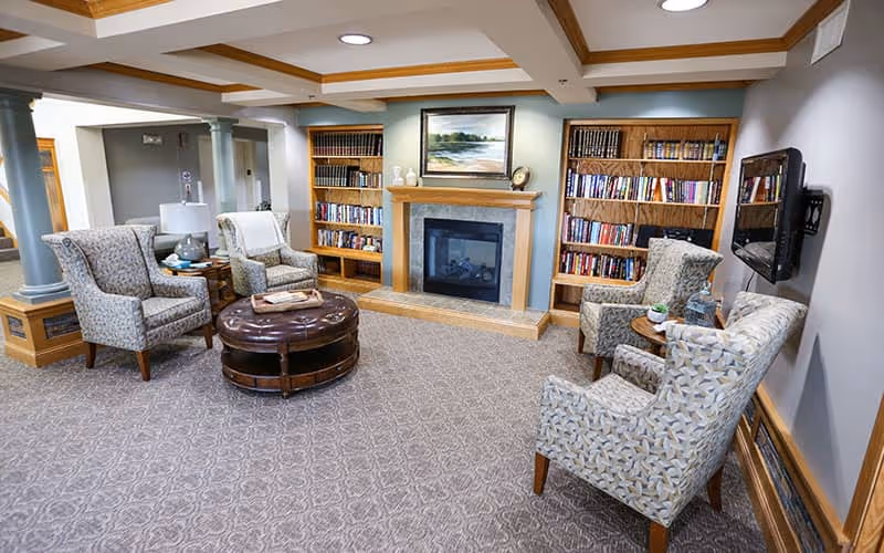 A cozy senior living common area with four patterned armchairs arranged around a round leather ottoman. The room features a fireplace with a wooden mantel, flanked by two built-in bookshelves filled with books. A flat-screen TV is mounted on the wall to the right. The ceiling has wooden beams, and the carpet has a subtle geometric pattern.