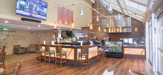 Interior view of a modern dining and bar area with wooden floors, a bar counter with high chairs, a display case with food items, and a large TV mounted on the wall. The space is well-lit with natural light coming through large windows and ceiling skylights. There are additional seating areas with armchairs in the background.