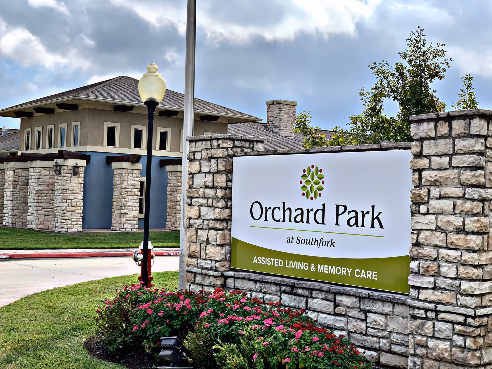 Stone entrance sign for Orchard Park at Southfork Assisted Living & Memory Care with a landscaped flower bed and a building in the background under a cloudy sky.