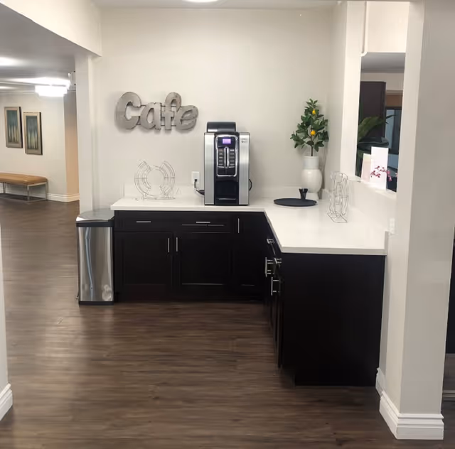 A small cafe area in a senior living facility with a coffee machine on a white countertop, dark wood cabinets below, a metal trash can to the left, and a decorative plant in a white vase on the right. The word 'Cafe' is mounted on the wall above the coffee machine. The floor is wood, and there is a hallway with framed pictures and a bench visible in the background.