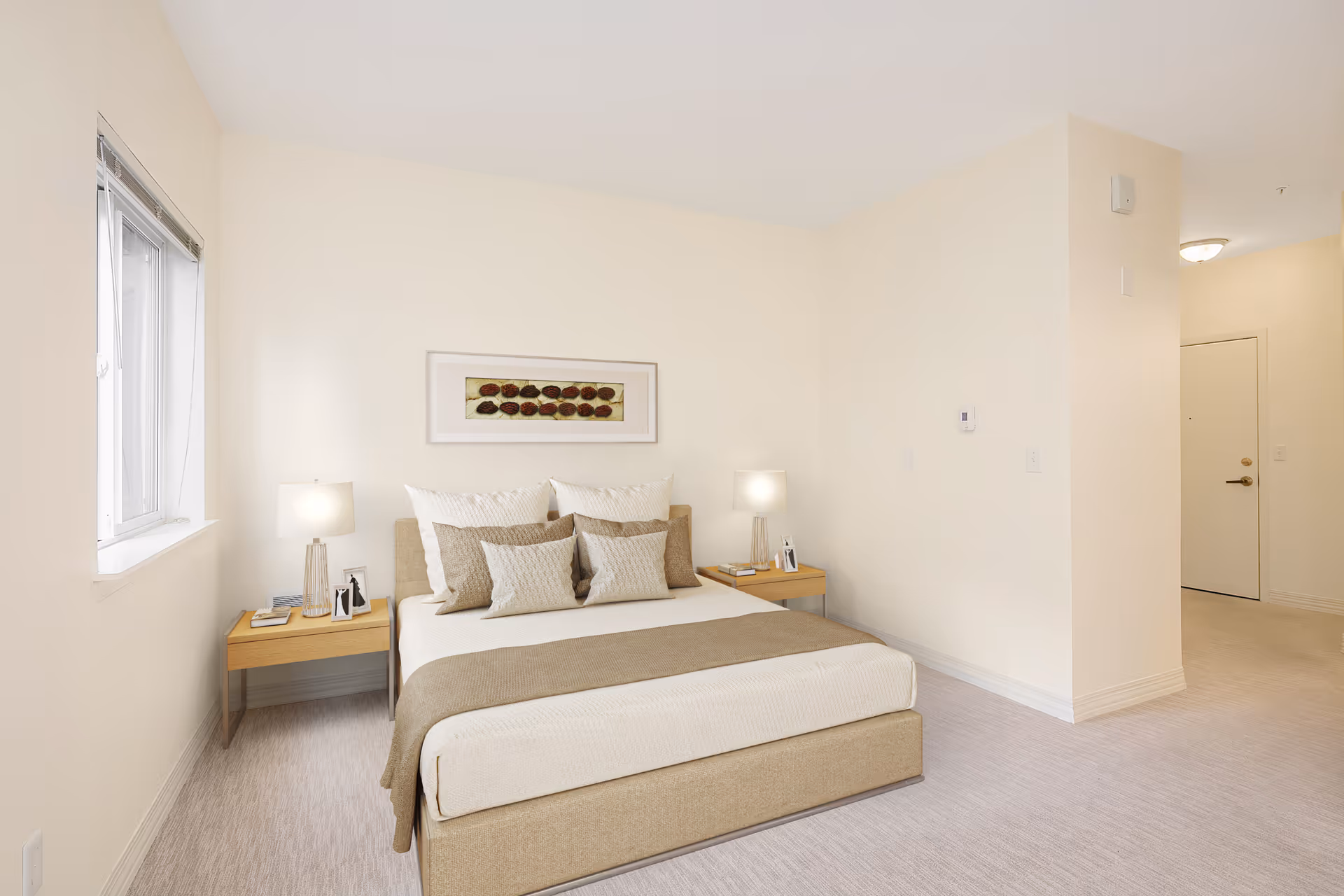 A neatly arranged bedroom with a beige upholstered bed featuring multiple pillows and a beige throw blanket. There are two wooden nightstands on either side of the bed, each with a lamp and small decorative items. A framed artwork hangs above the bed. The room has light-colored walls and carpet, a window on the left side, and a door visible in the background.