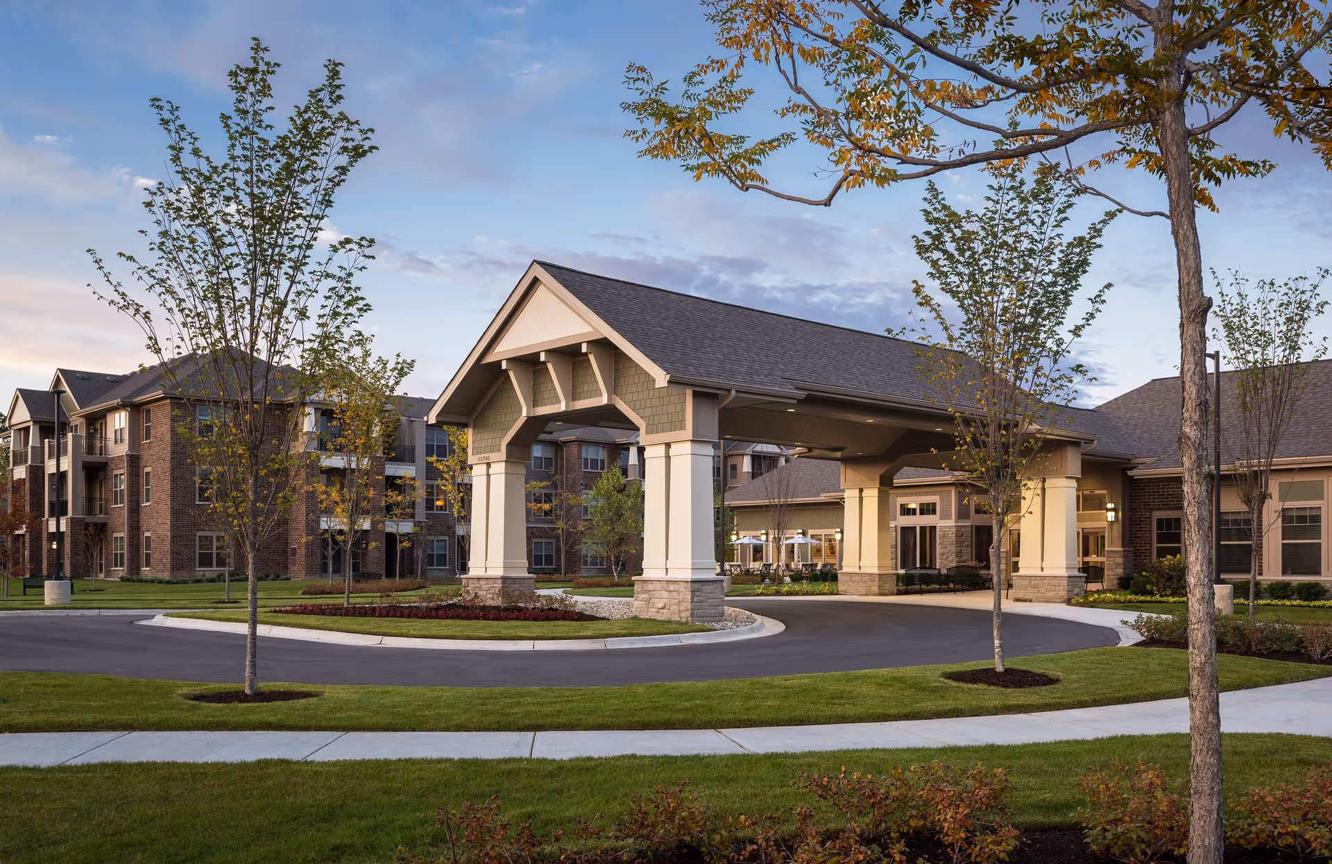 Exterior view of Cedarbrook of Northville senior living facility at dusk, showing a covered entrance with pillars, landscaped grounds with trees and grass, and multi-story residential buildings in the background.