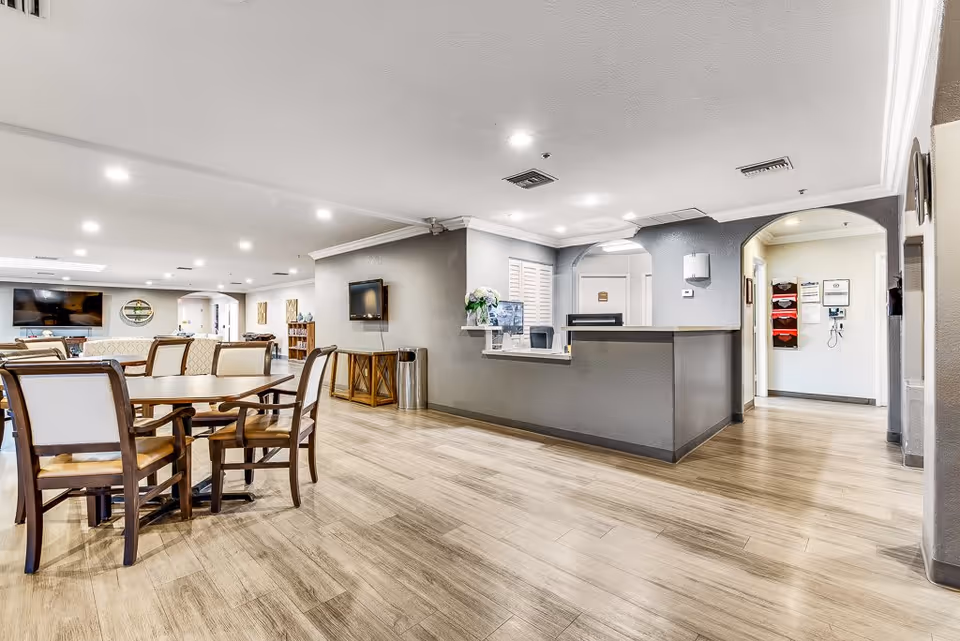 A spacious and well-lit senior living facility common area with wooden flooring, several wooden chairs and tables, a reception desk, wall-mounted TV, and decorative elements including flowers and artwork. The area has a modern and clean design with recessed lighting and an open layout.
