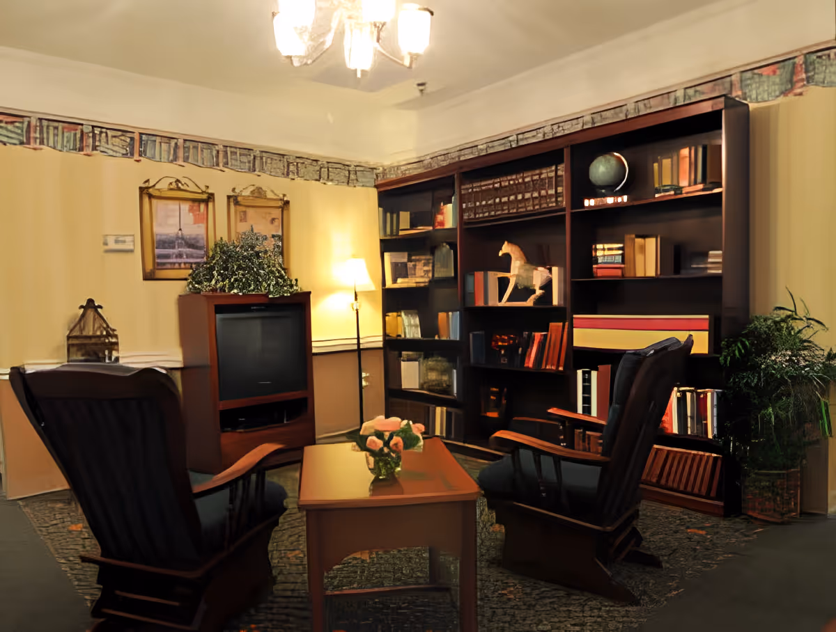 A cozy living room with two dark wooden rocking chairs facing a wooden coffee table with a vase of flowers. Behind the chairs is a large bookshelf filled with books and decorative items, including a globe and a wooden horse. A TV sits on a wooden stand with a plant on top, and a floor lamp provides warm lighting. The walls are decorated with framed pictures and a wallpaper border near the ceiling.