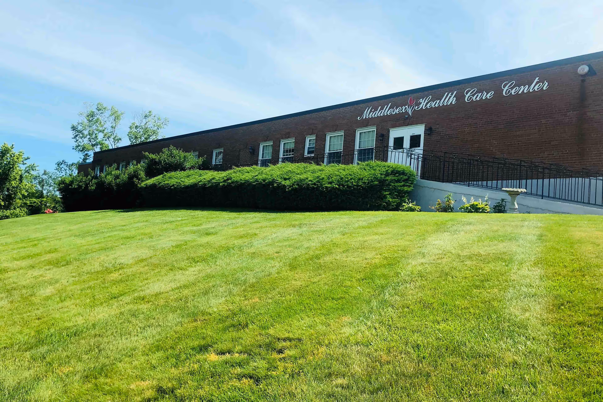 Exterior view of Middlesex Health Care Center, a brick building with several windows and a white entrance door. The building is surrounded by a well-maintained green lawn and bushes under a clear blue sky.