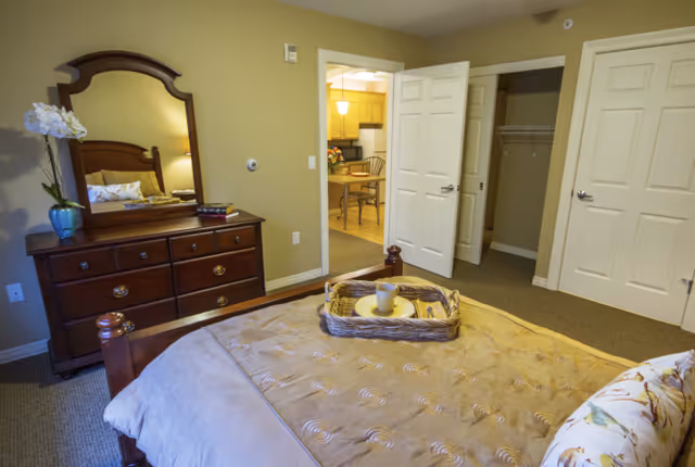 A cozy bedroom in an assisted living community featuring a wooden bed with a beige comforter and floral pillow, a wooden dresser with a mirror and a vase of white flowers, an open door leading to a kitchen and another door to a closet.