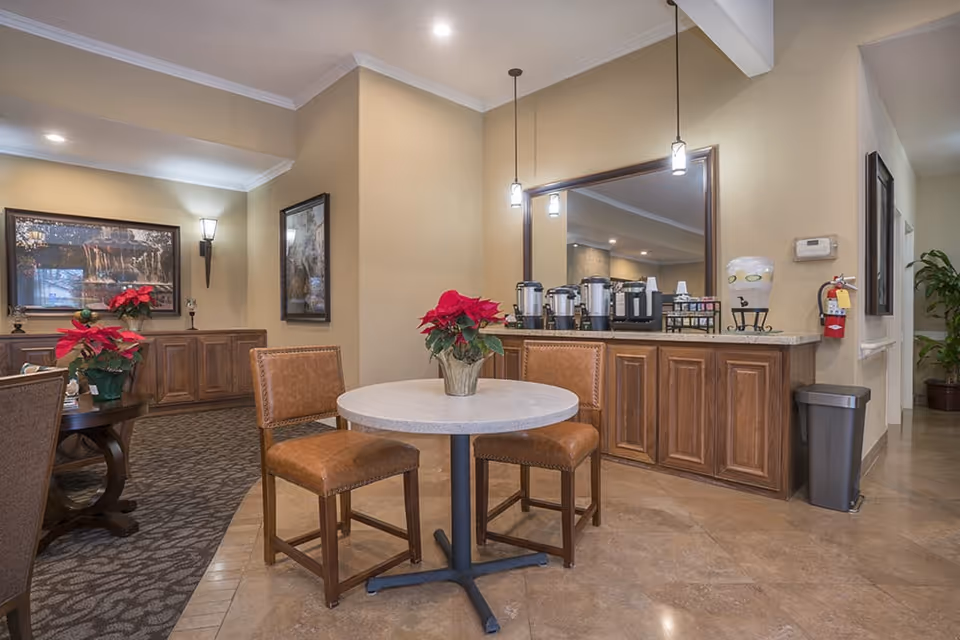 A cozy senior living common area with a small round table and two brown leather chairs. The table has a red poinsettia plant in the center. Behind the table is a counter with coffee dispensers, cups, and a water dispenser. The walls are beige with framed artwork and soft lighting from wall sconces and hanging pendant lights. There is a carpeted seating area with more poinsettia plants and wooden cabinets along the wall.