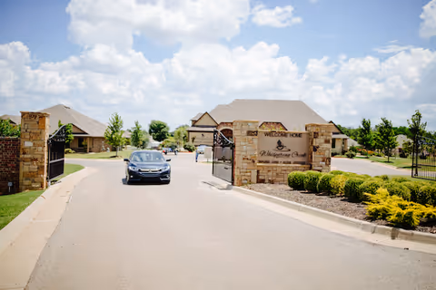 Entrance to Whispering Creek senior living facility with a car driving on the road. Stone pillars and a sign that reads 'Welcome Home Whispering Creek' are visible, along with gated entry and landscaped greenery under a partly cloudy sky.