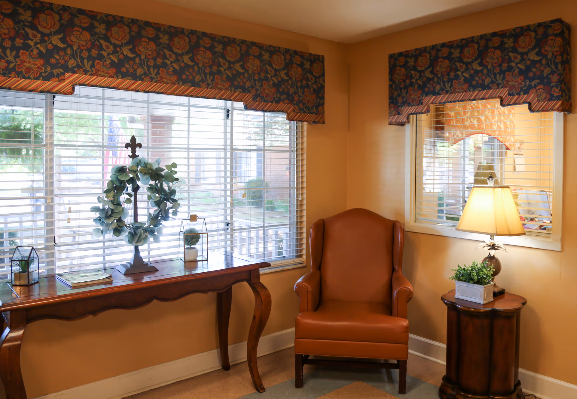 A cozy corner in a living room with a brown leather armchair next to a small wooden side table holding a lamp and a small potted plant. Behind the chair is a window with white blinds and a floral valance. To the left is a wooden console table with decorative items including a wreath and small plants, positioned in front of a large window with white blinds and a matching floral valance.