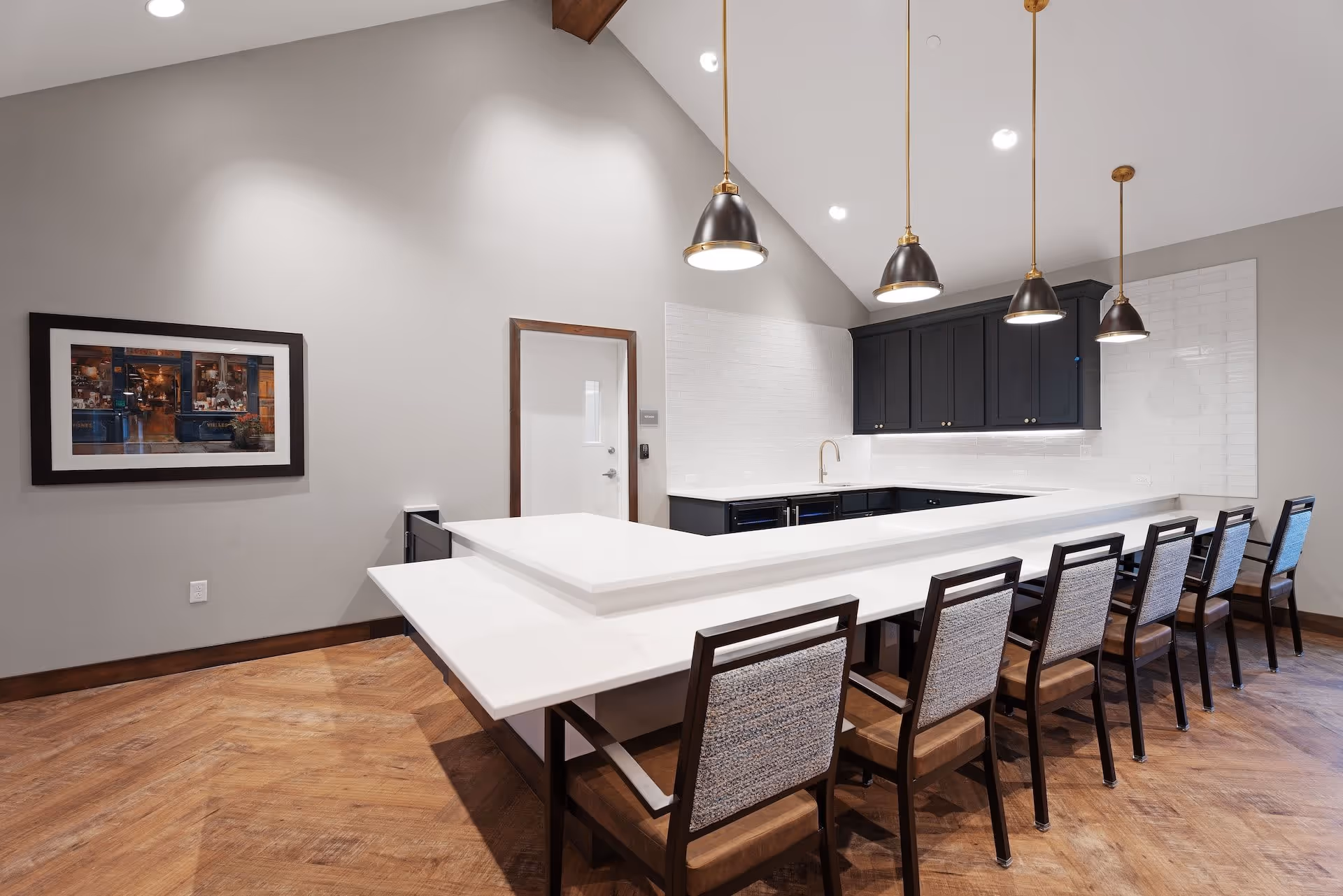 Modern kitchen area with a long white countertop bar and seven chairs with wooden frames and cushioned seats. The kitchen features black cabinets, a white tiled backsplash, and four pendant lights hanging from a vaulted ceiling. A framed picture hangs on the light gray wall, and the floor is a warm wood pattern.