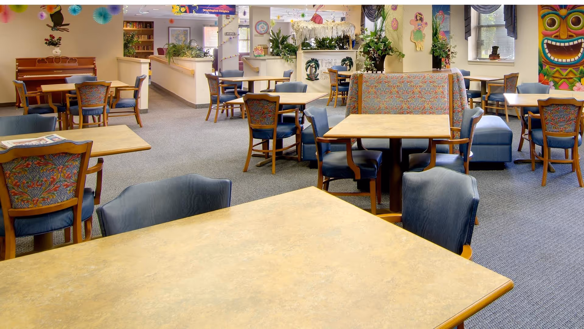 A spacious dining area in a senior living facility with multiple tables and chairs arranged neatly. The room features colorful patterned upholstery on chairs and a bench, a piano in the corner, decorative wall art including a tiki mask and a fairy, and various plants and decorations throughout the space.