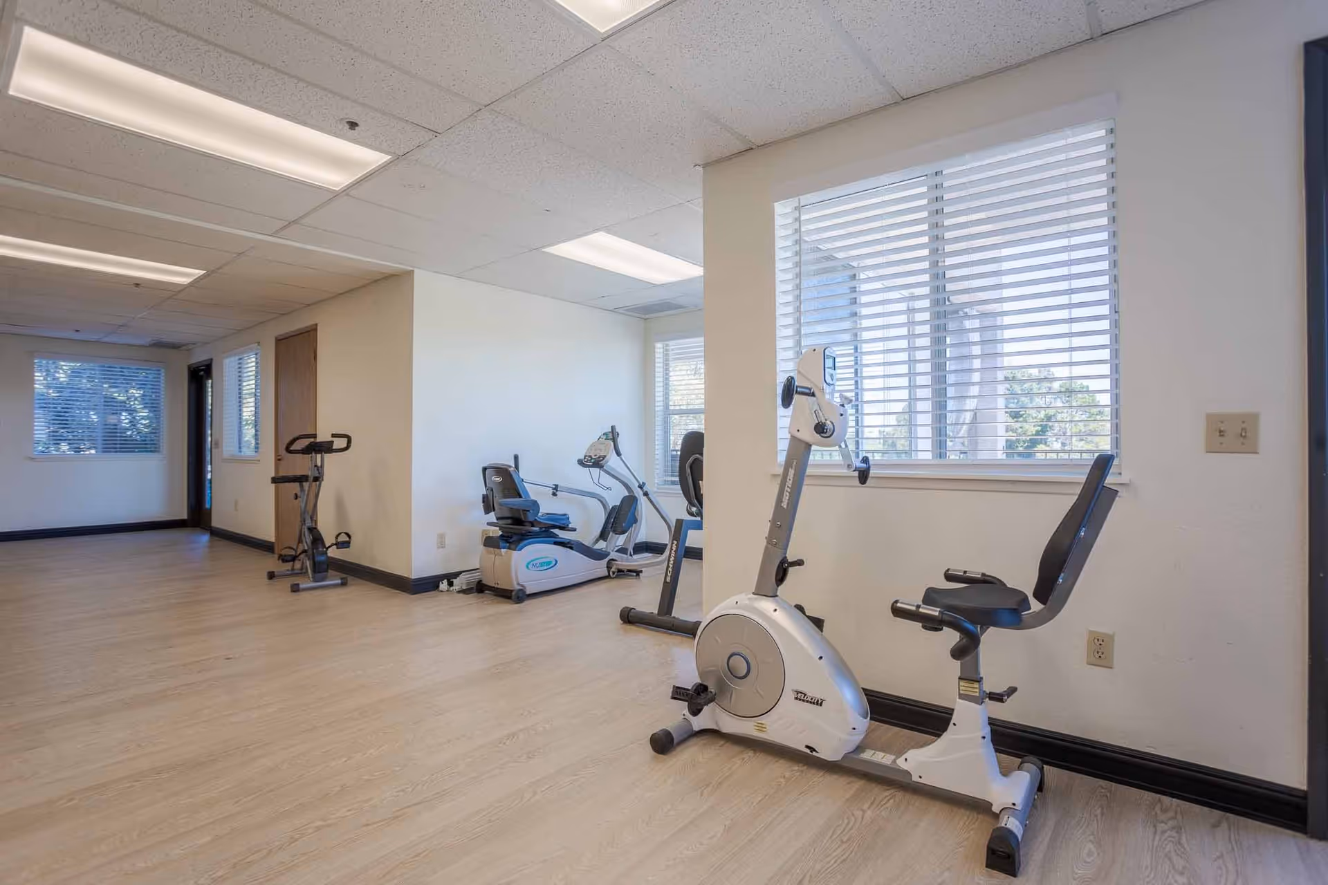 A small exercise room with light wood flooring and white walls, featuring three stationary exercise bikes positioned near windows with white blinds. The room is well-lit with fluorescent ceiling lights.