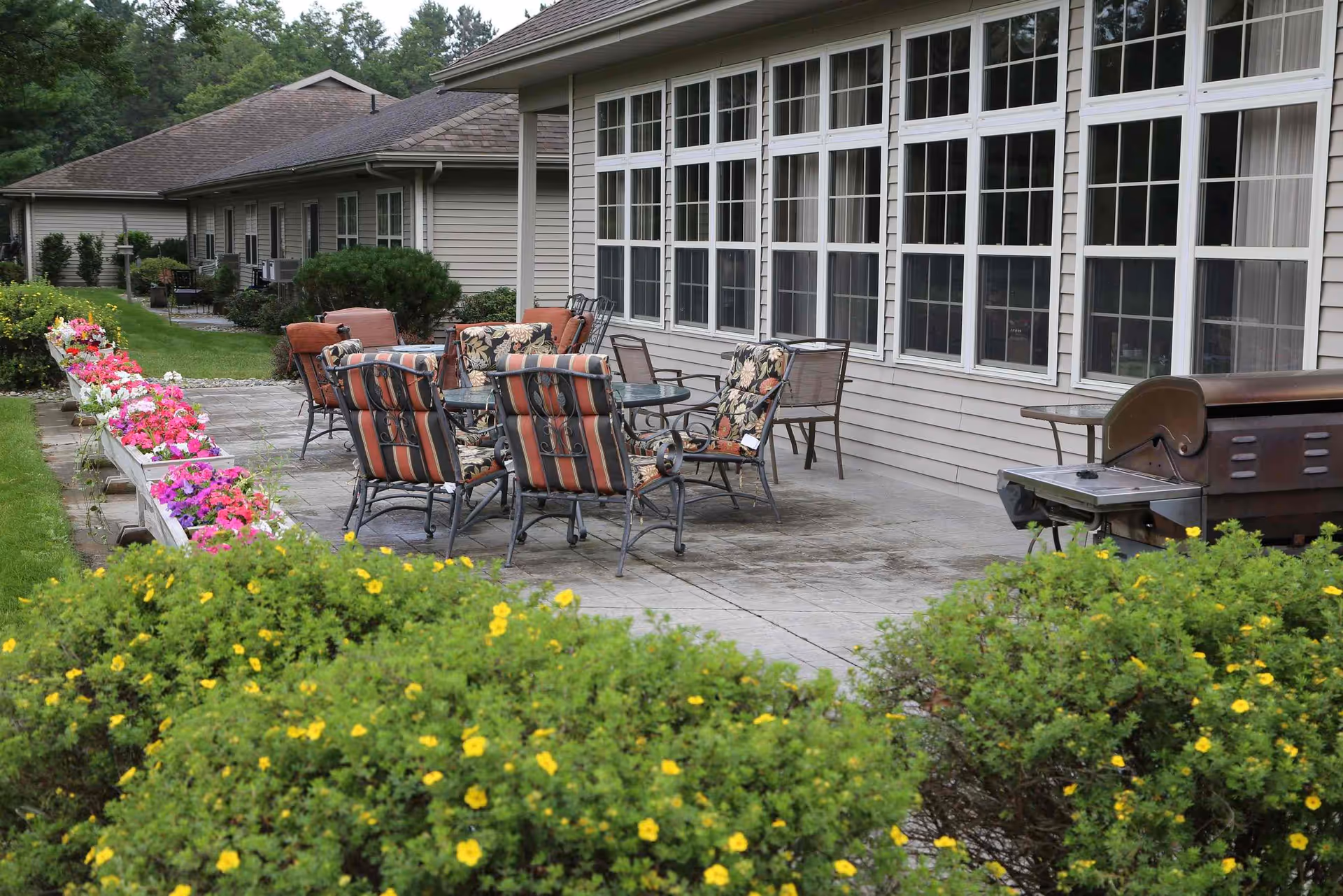 Outdoor patio with a table, cushioned chairs, a grill, and flower beds alongside a building with large windows.