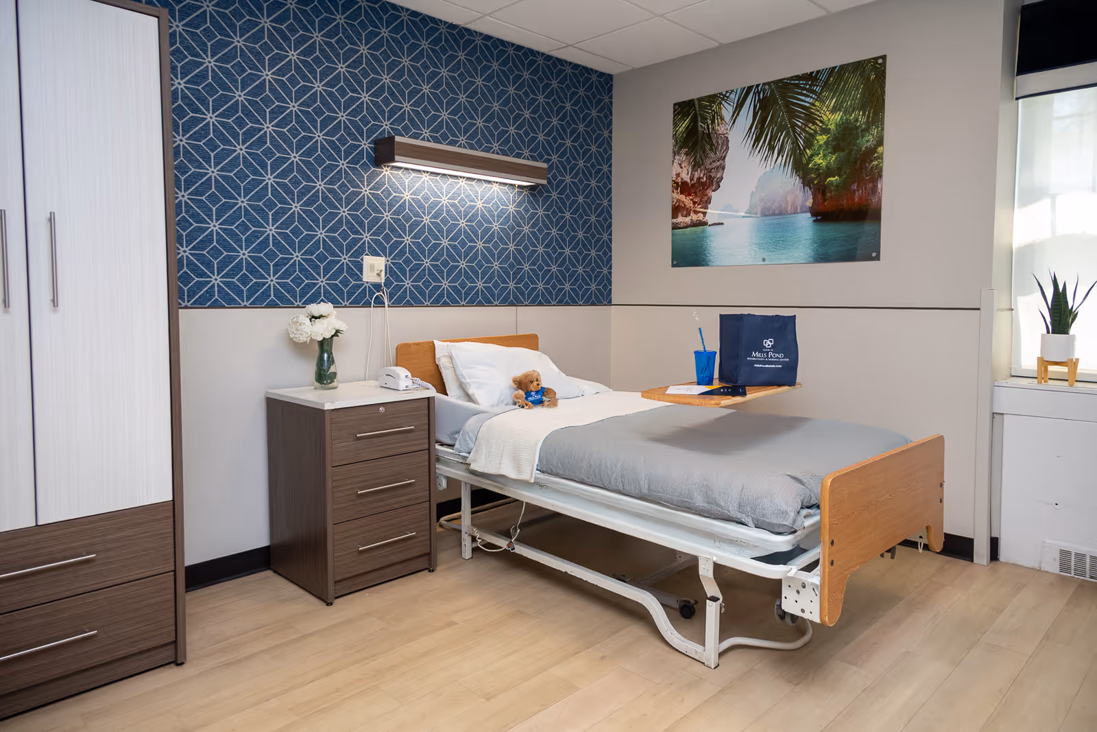 Bright nursing facility bedroom with a hospital bed, bedside table and wardrobe under patterned wallpaper and tropical wall art.