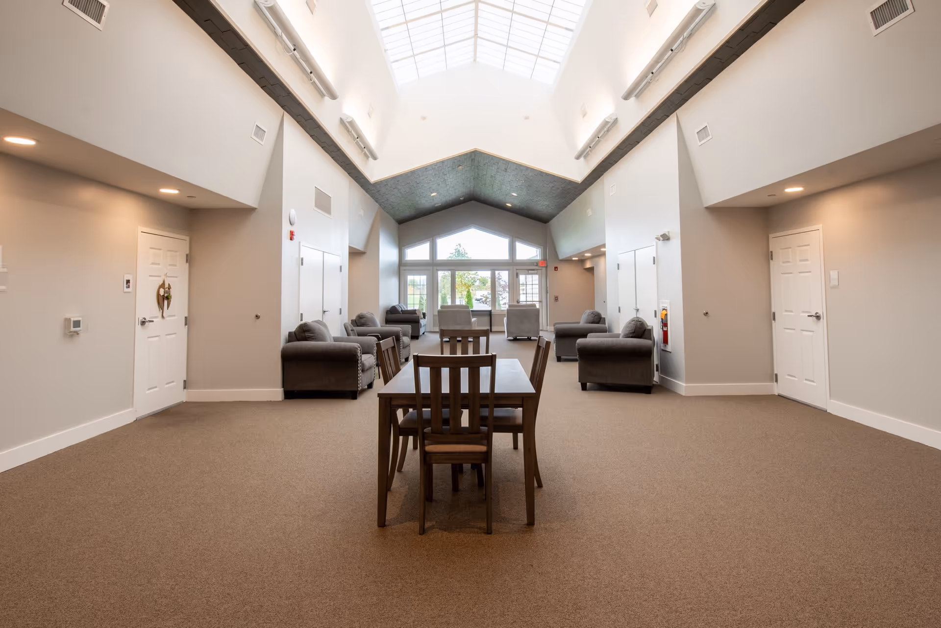 A spacious, well-lit common area with a high ceiling and skylight. The room features a wooden table with four chairs in the center, surrounded by several gray upholstered armchairs arranged along the walls. Large windows at the far end provide natural light and a view of greenery outside. The walls are painted light beige, and there are multiple doors and vents visible.