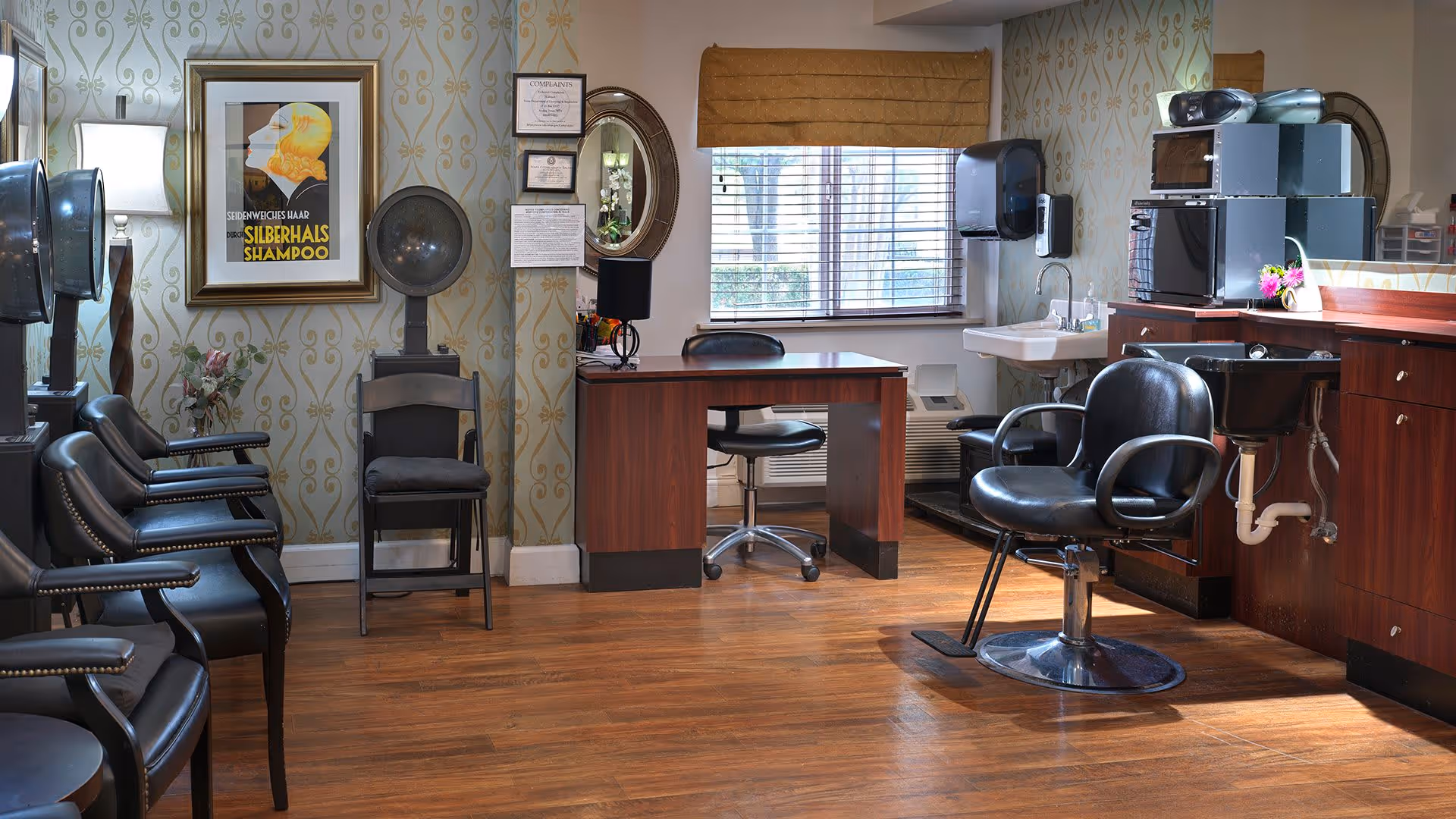 Interior of a hair salon room with wooden flooring, black salon chairs, hair dryers, a wooden desk with a black chair, a window with blinds and a brown valance, a sink, and a microwave on a countertop. The walls have patterned wallpaper and framed artwork.