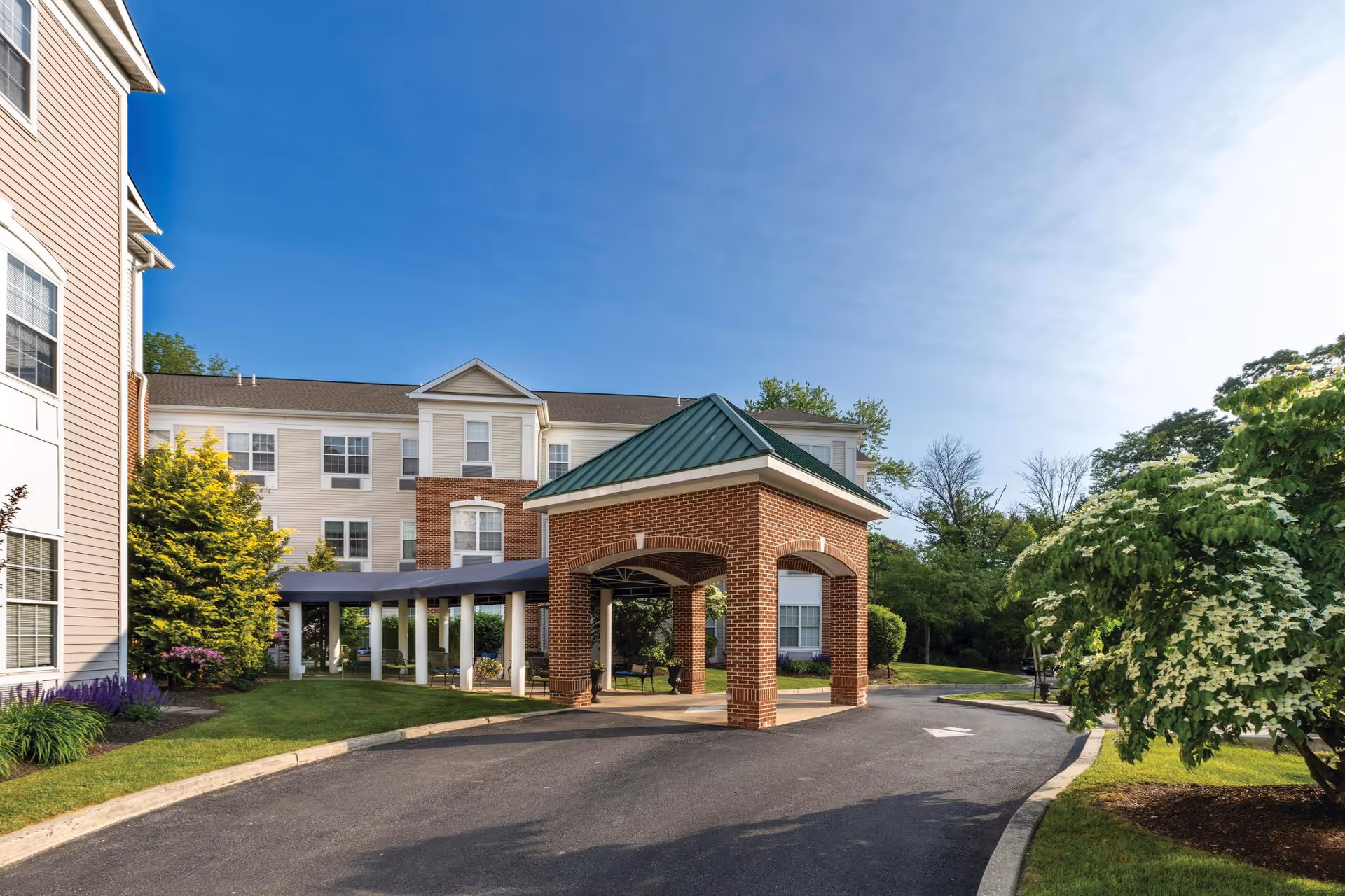 Exterior view of Rose Tree Place senior living facility showing a driveway with a covered entrance supported by brick columns, surrounded by well-maintained landscaping and trees under a clear blue sky.
