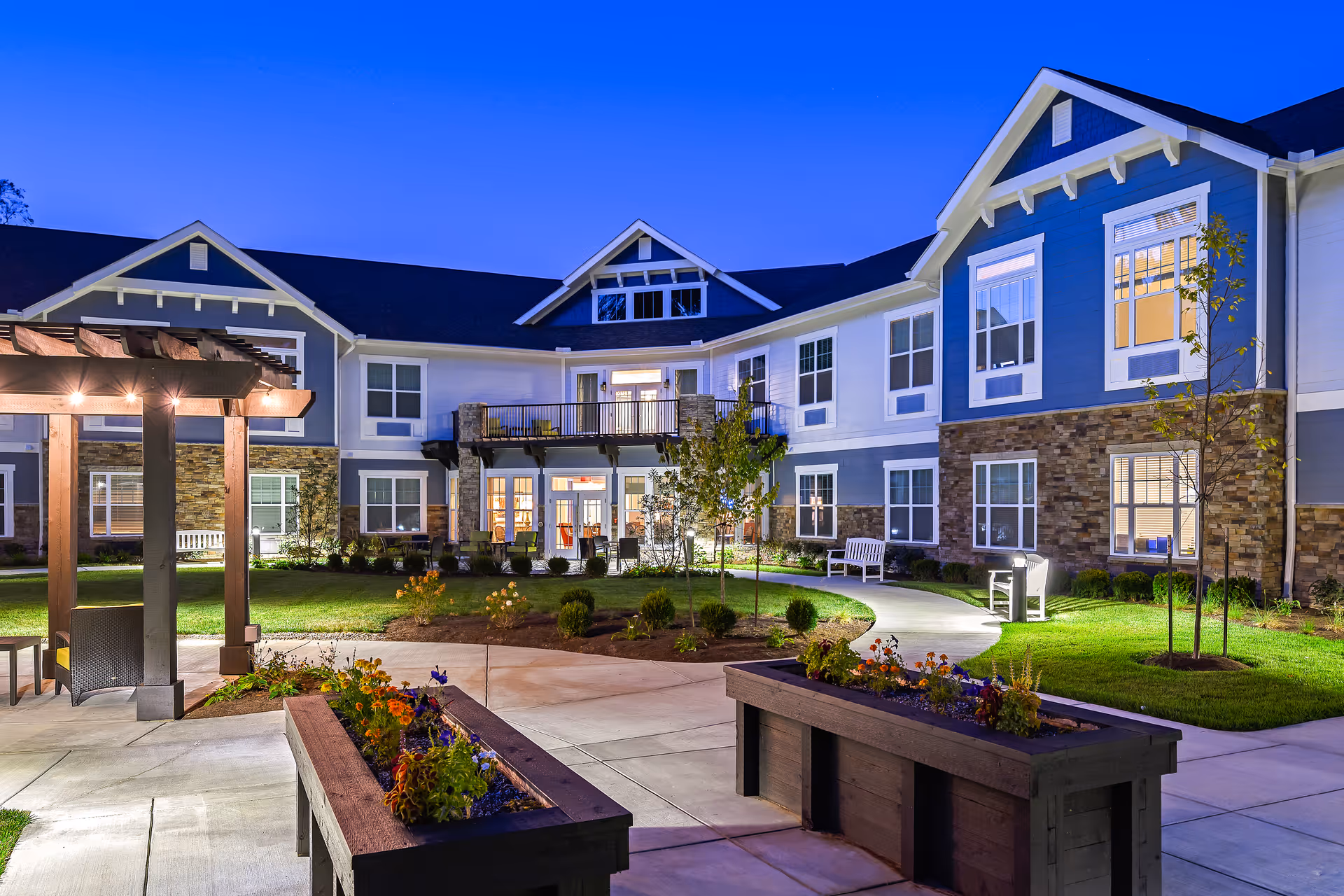 Evening view of the exterior courtyard of The Ashton at Anderson senior living facility, featuring a well-lit two-story building with blue and white siding, stone accents, a pergola with lights, benches, landscaped garden beds, and a curved concrete walkway.