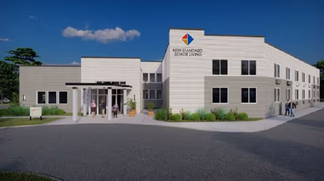 Exterior view of New Standard Senior Living at Egg Harbor Township, showing a modern two-story building with a light-colored facade, several windows, and a covered entrance with people walking nearby under a clear blue sky.