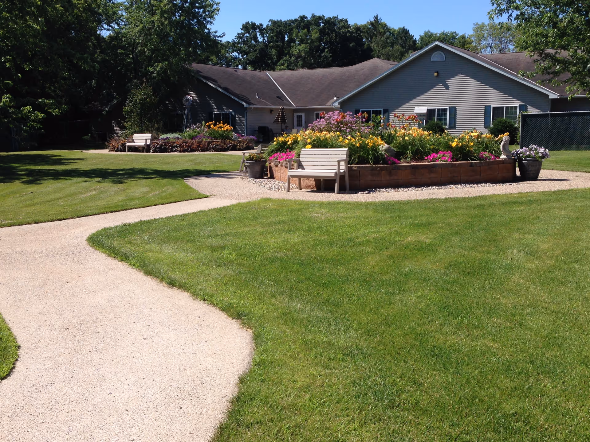 A well-maintained outdoor garden area with a paved walkway, green grass, and flower beds filled with colorful flowers. There are white benches placed around the flower beds, and a single-story building with gray siding and multiple windows is visible in the background under a clear blue sky.