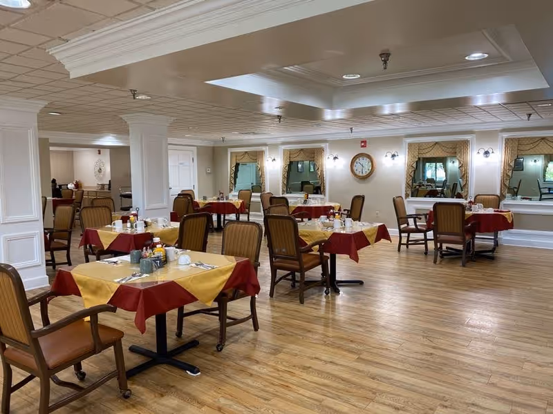 A spacious dining room with several tables covered in red and yellow tablecloths. Each table is set with cups, plates, and condiments. The room has wooden flooring, beige walls, large mirrors, and a clock on the wall. Comfortable wooden chairs with cushions are arranged around the tables.