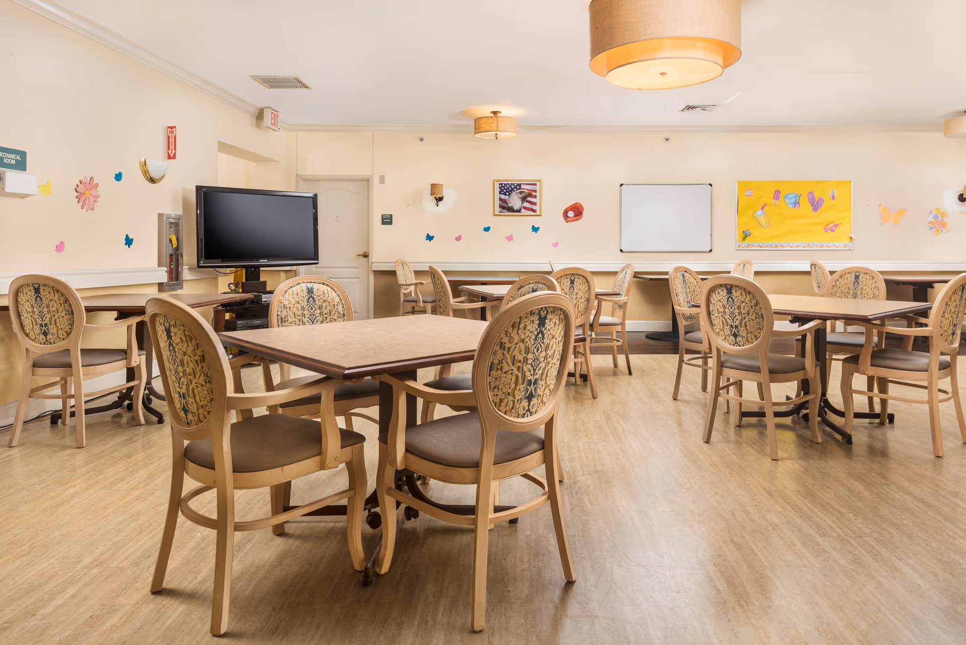 A bright, clean dining room with multiple square tables and cushioned chairs arranged neatly. The walls are decorated with colorful paper cutouts and bulletin boards, and a large flat-screen TV is mounted on one wall. The room has wooden flooring and ceiling lights with beige lampshades.
