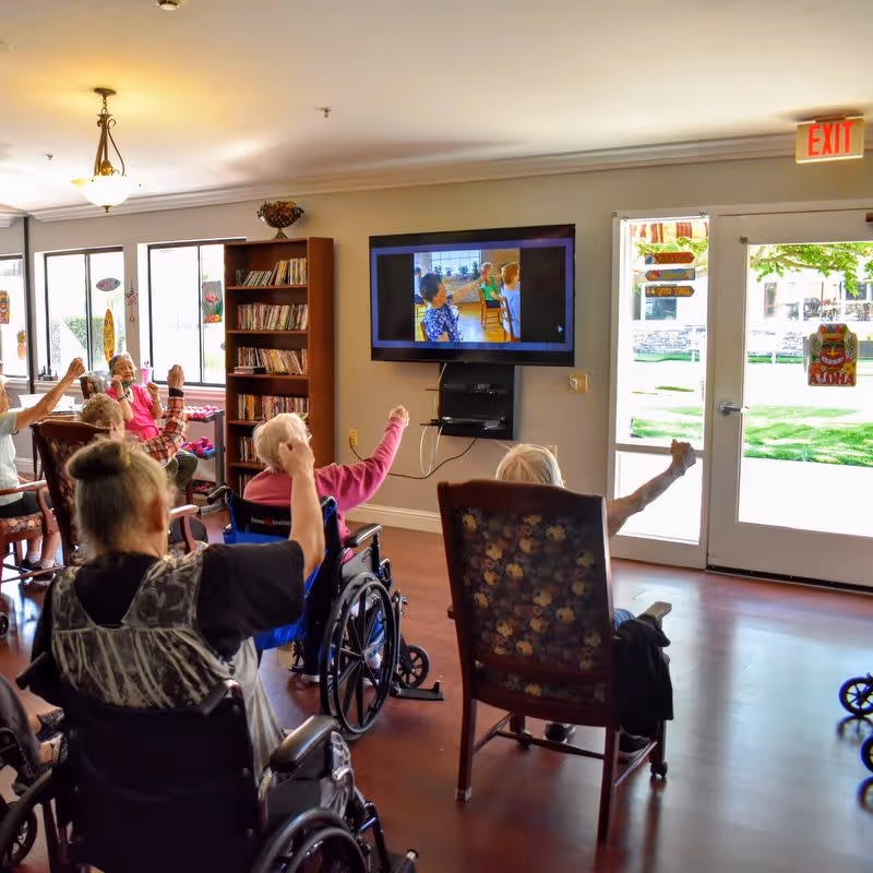 Residents in wheelchairs and chairs raising their arms while watching a wall-mounted TV in a bright communal room.