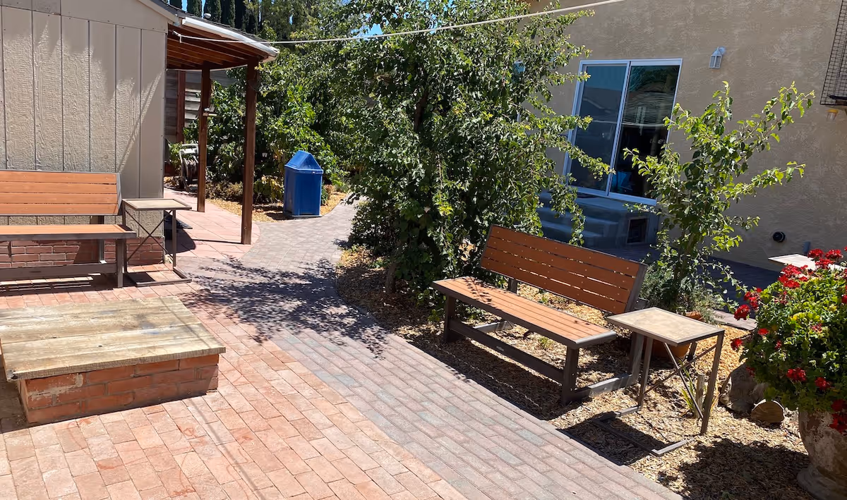 Outdoor patio area with brick flooring, two wooden benches with metal frames, a small side table, green leafy bushes, a large planter with red flowers, and a building with a window in the background.