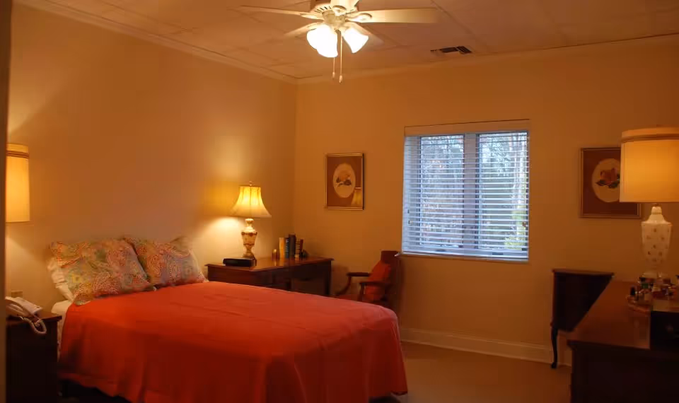 A cozy bedroom with a double bed covered in a red bedspread and colorful pillows. The room has warm lighting from two table lamps on wooden nightstands, a ceiling fan with lights, a window with blinds, and framed artwork on the walls.
