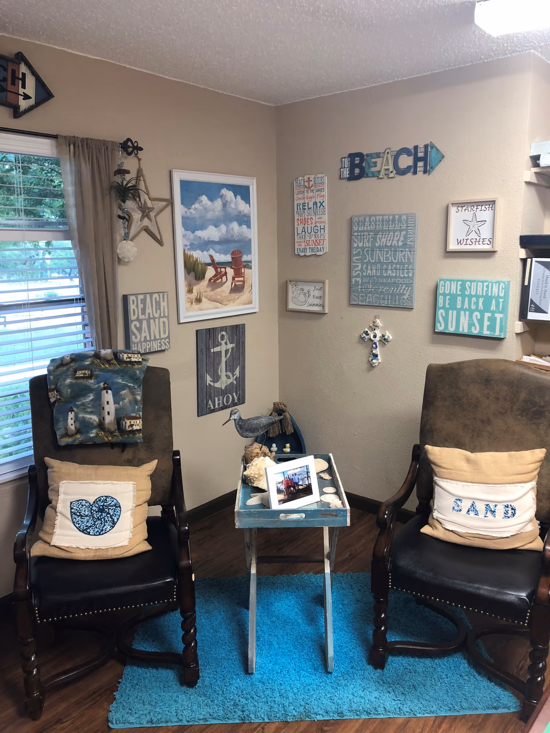 A cozy corner with two dark wooden armchairs, each with a beige pillow, one with a heart design and the other with the word 'SAND'. Between the chairs is a small blue tray table holding seashells and a framed photo. The walls are decorated with beach-themed signs and artwork, including a painting of two chairs on a sandy beach, an anchor sign, and various inspirational quotes. A window with beige curtains is on the left, and the floor has a bright blue rug.