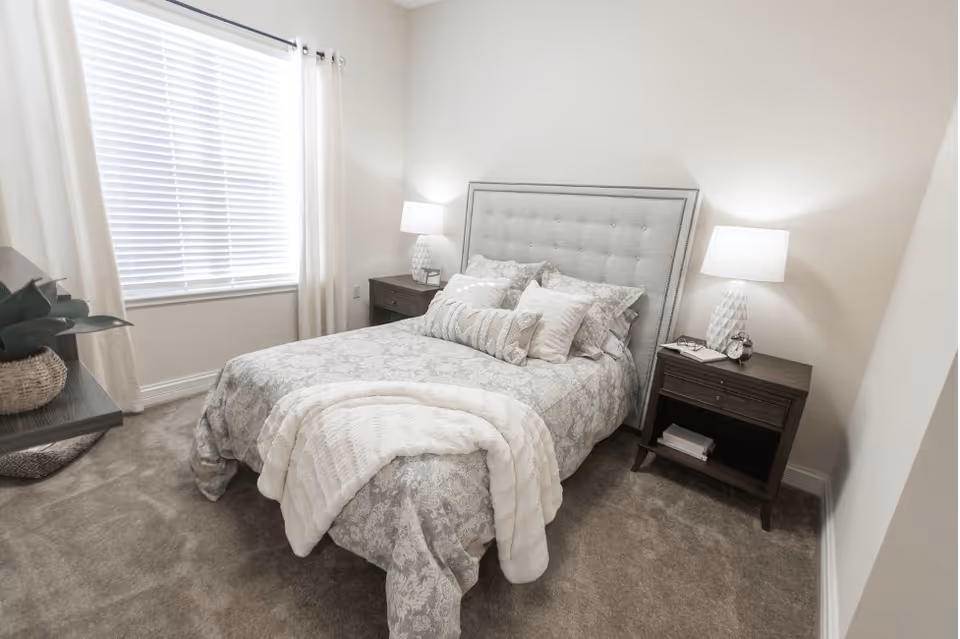 Neutral-toned bedroom with a neatly made bed, upholstered headboard, two bedside tables with lamps, and a window with blinds.