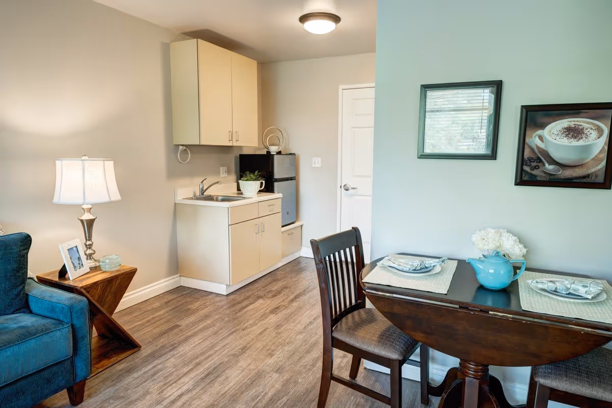 A cozy interior space featuring a small kitchenette with a sink, cabinets, and a mini refrigerator. Adjacent to the kitchenette is a dark wooden dining table set for two with placemats, plates, and a blue teapot with white flowers. A blue upholstered chair and a wooden side table with a lamp and a framed photo are visible on the left. The walls are light-colored with two framed pictures hanging above the dining table.